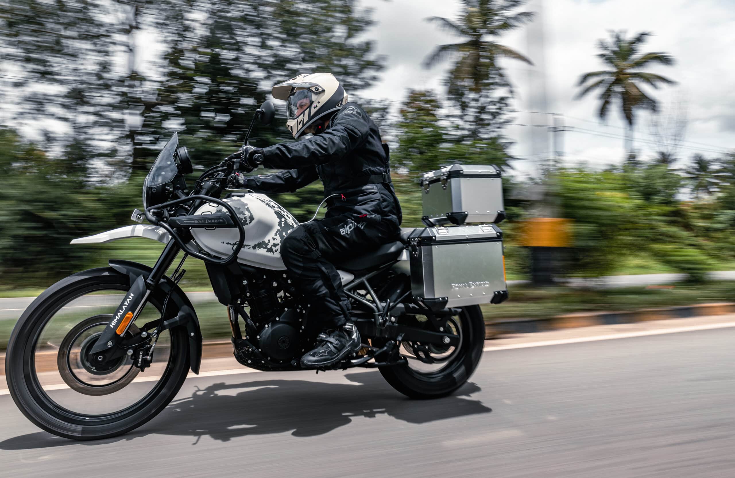 A rider in full gear on a white Royal Enfield Himalayan motorcycle with silver panniers, riding on a road with palm trees in the background.
