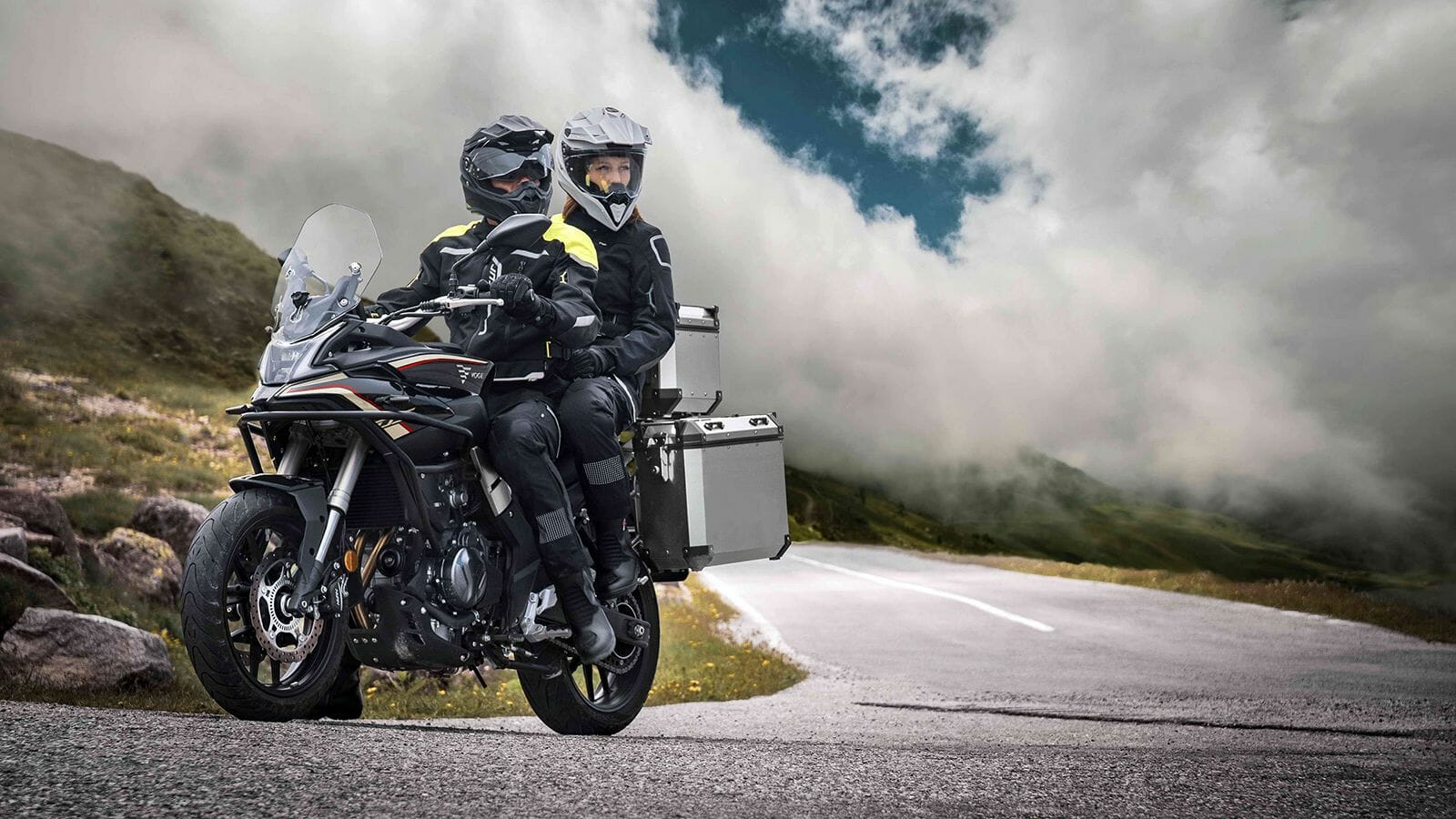 A couple in protective gear riding a black adventure motorcycle with aluminum panniers on a scenic mountain road under a cloudy sky in Greece.