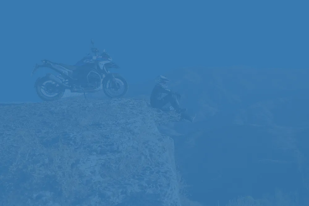 A motorcyclist in riding gear sits on a cliff edge next to their adventure motorcycle, enjoying a scenic view during a tour in Greece or the Balkans.