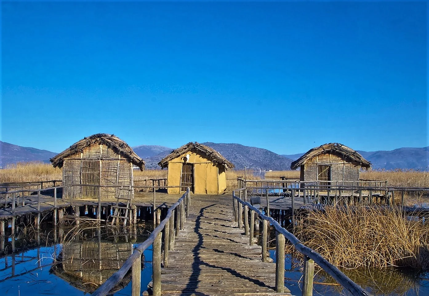 Traditional wooden huts on stilts connected by a wooden walkway over a calm lake, set against a clear blue sky and distant mountains in Greece.