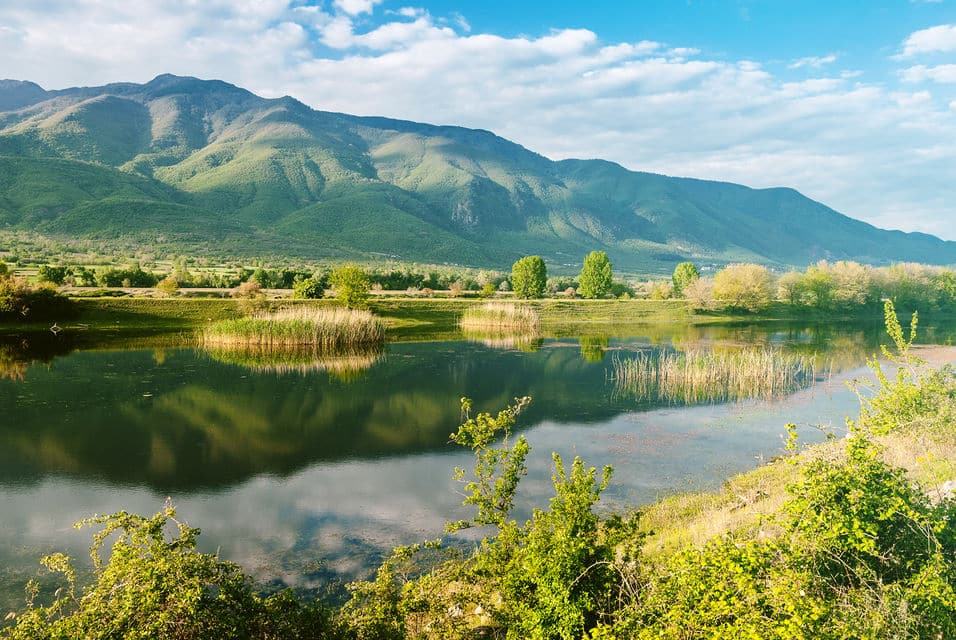 A serene lake reflecting green mountains and blue skies, surrounded by lush vegetation, perfect for scenic motorcycle touring in the Greek countryside.