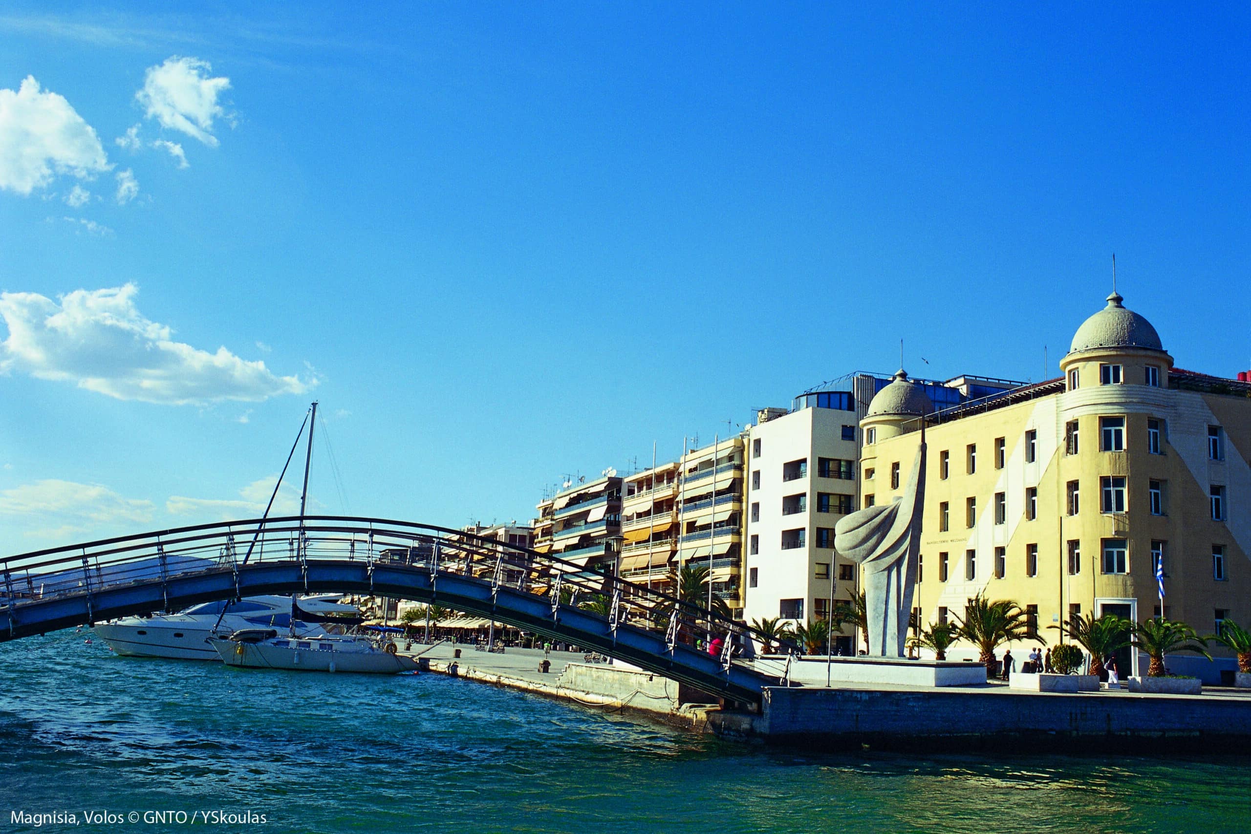 A scenic view of the waterfront in Volos, Greece, featuring a curved pedestrian bridge, historic buildings, and boats under a bright blue sky.