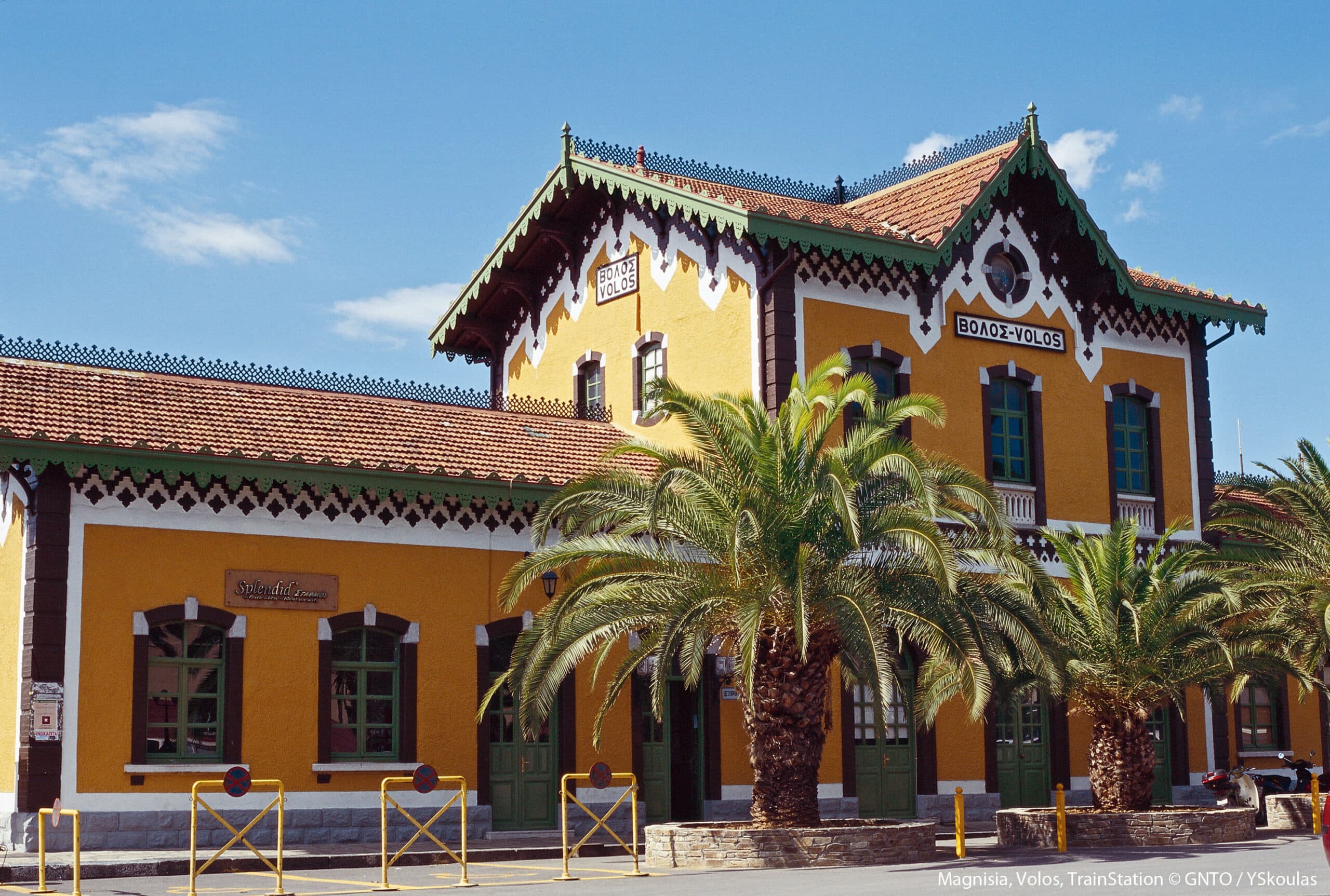 The historic yellow Volos train station building in Greece, featuring traditional architecture, green trim, and palm trees under a clear blue sky.