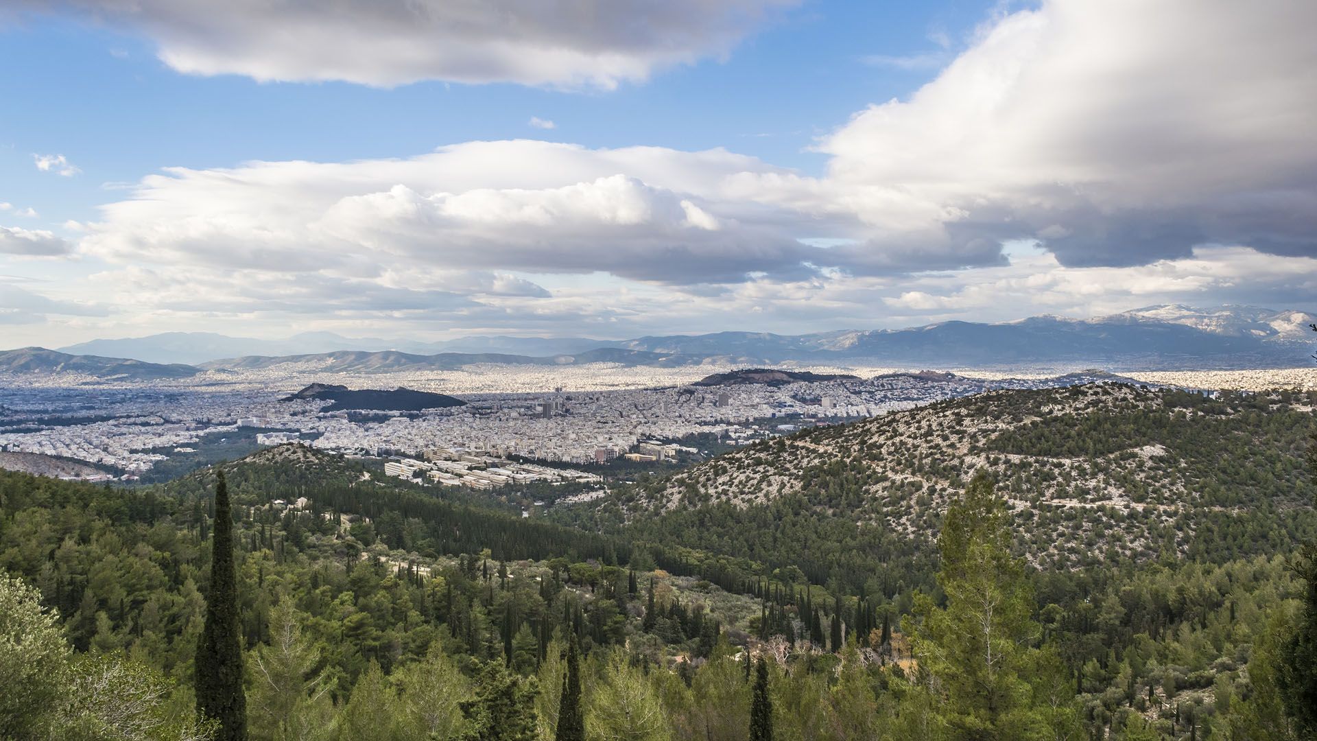 Πανοραμική άποψη της πόλης της Αθήνας που απλώνεται στον ορίζοντα, από μια καταπράσινη, πευκόφυτη πλαγιά κάτω από έναν φωτεινό ουρανό με διάσπαρτα σύννεφα.
