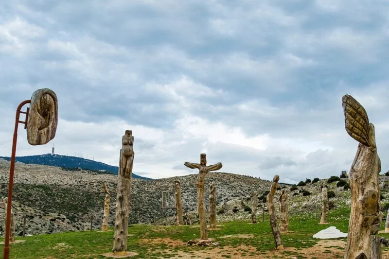 Ξύλινα γλυπτά στέκονται σε ένα καταπράσινο χωράφι ενάντια σε έναν συννεφιασμένο ουρανό, μια μοναδική στάση για ταξιδιώτες με μοτοσικλέτα που εξερευνούν τα γραφικά τοπία της Ελλάδας.