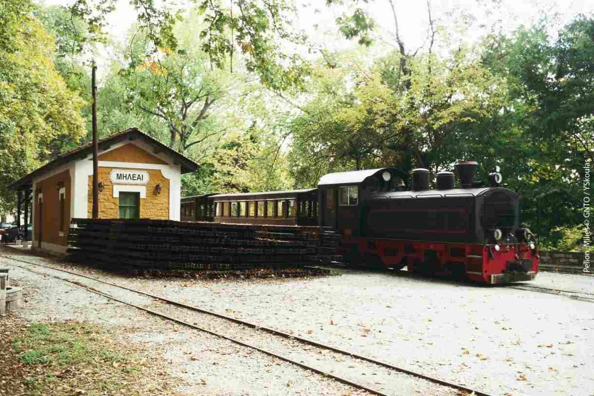 The historic steam train at the Milies station in Pelion, Greece, surrounded by lush trees, perfect for a scenic motorcycle adventure tour.