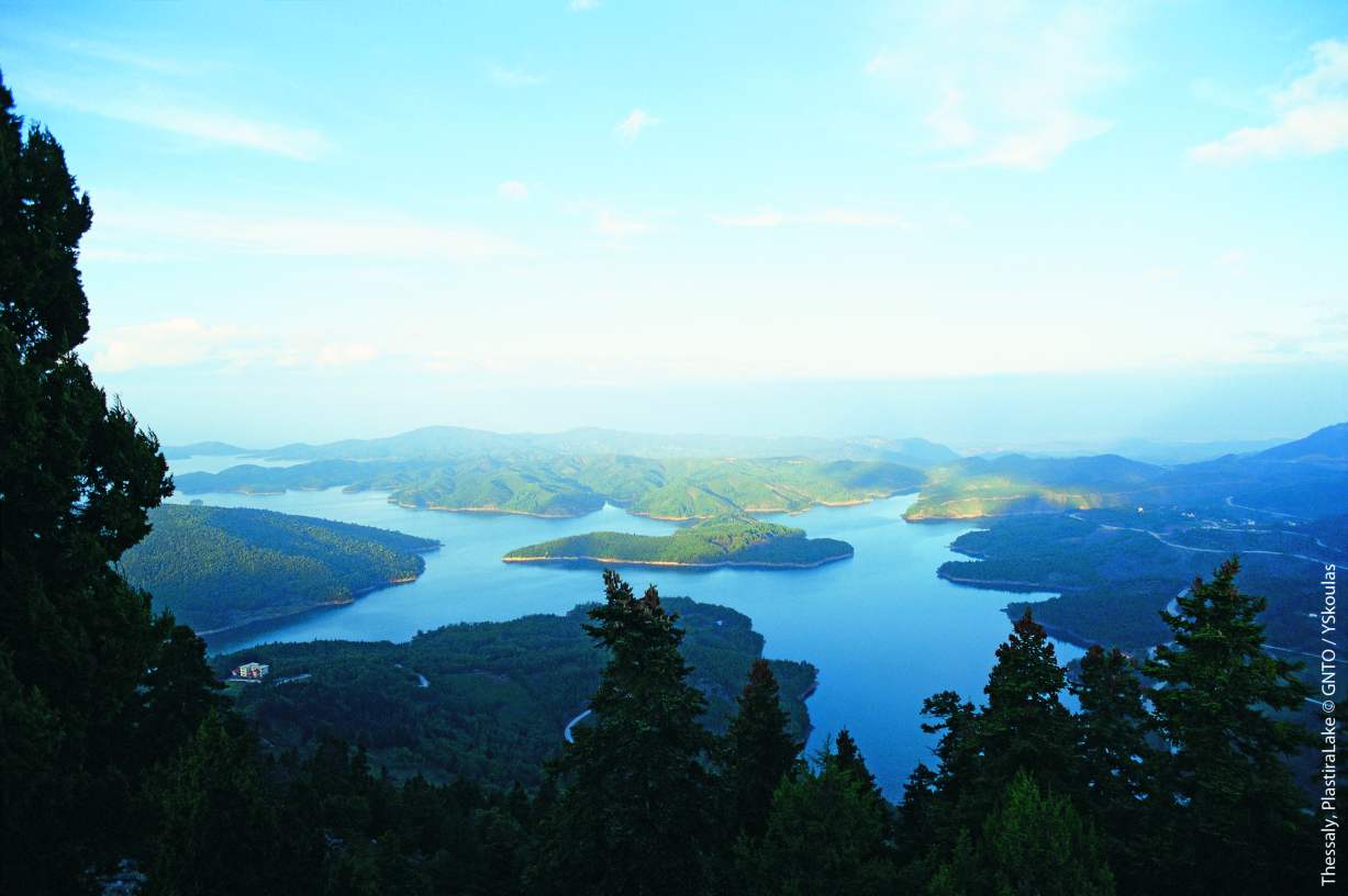 Panoramic view of Lake Plastiras in Thessaly, Greece, featuring blue water, forested islands, and surrounding mountains for motorcycle touring.
