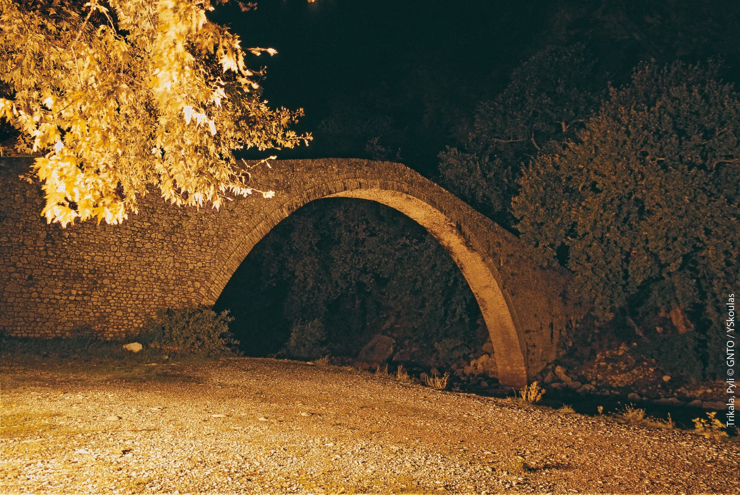 An ancient stone arched bridge in Pyli, Trikala, illuminated at night, surrounded by trees and a gravel riverbank during a motorcycle adventure.