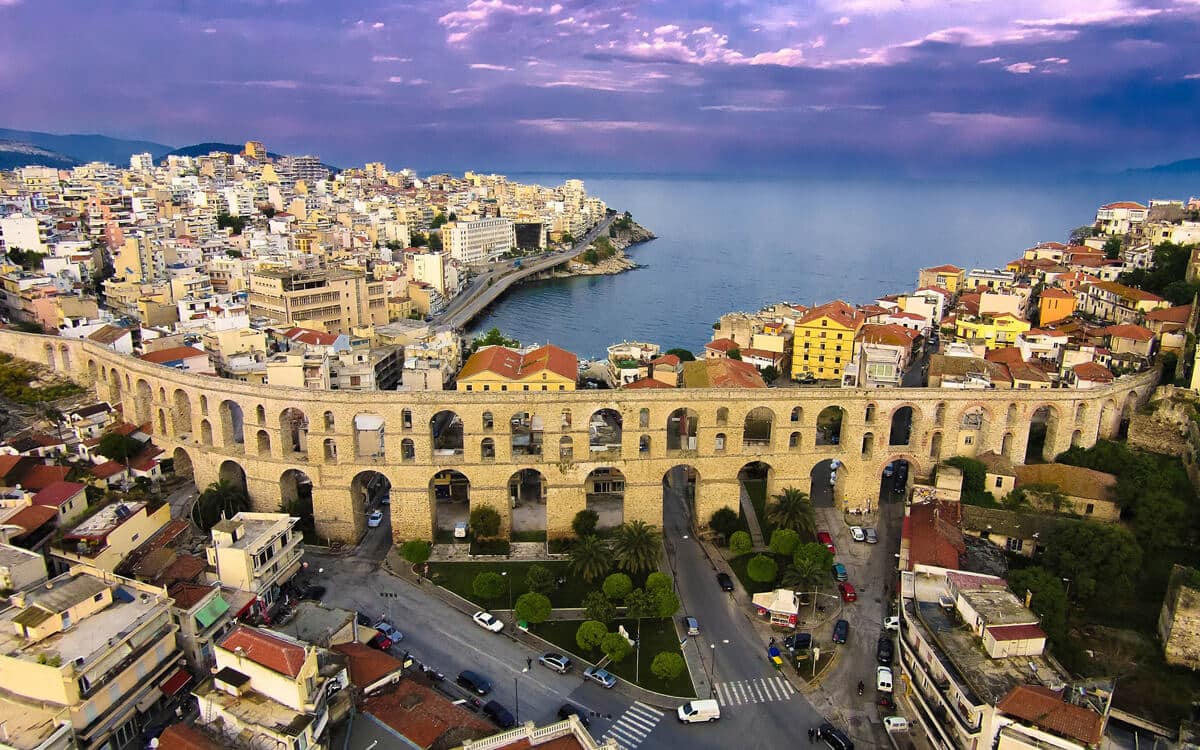 Aerial view of the historic Kamares aqueduct in Kavala, Greece, featuring stone arches overlooking the coastal city and the Aegean Sea at sunset.