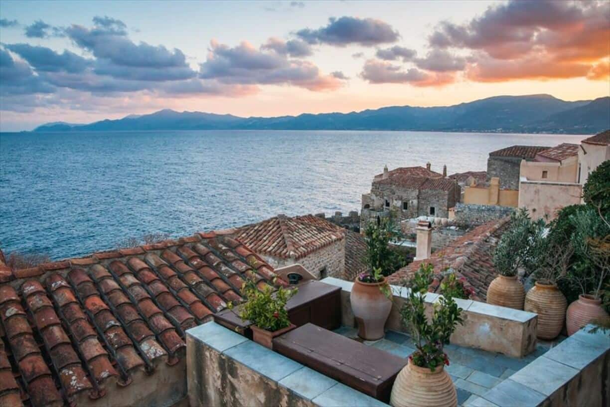 Scenic view of the historic Monemvasia castle town in Greece, featuring traditional tiled roofs, stone buildings, and the sea at sunset.