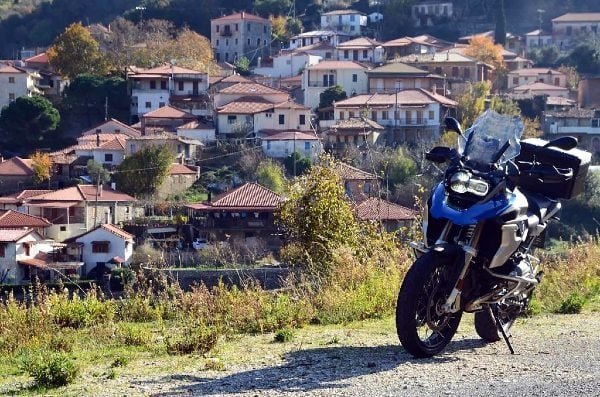A blue and silver adventure motorcycle parked on a gravel road with a traditional Greek mountain village in the background during a motorbike tour.