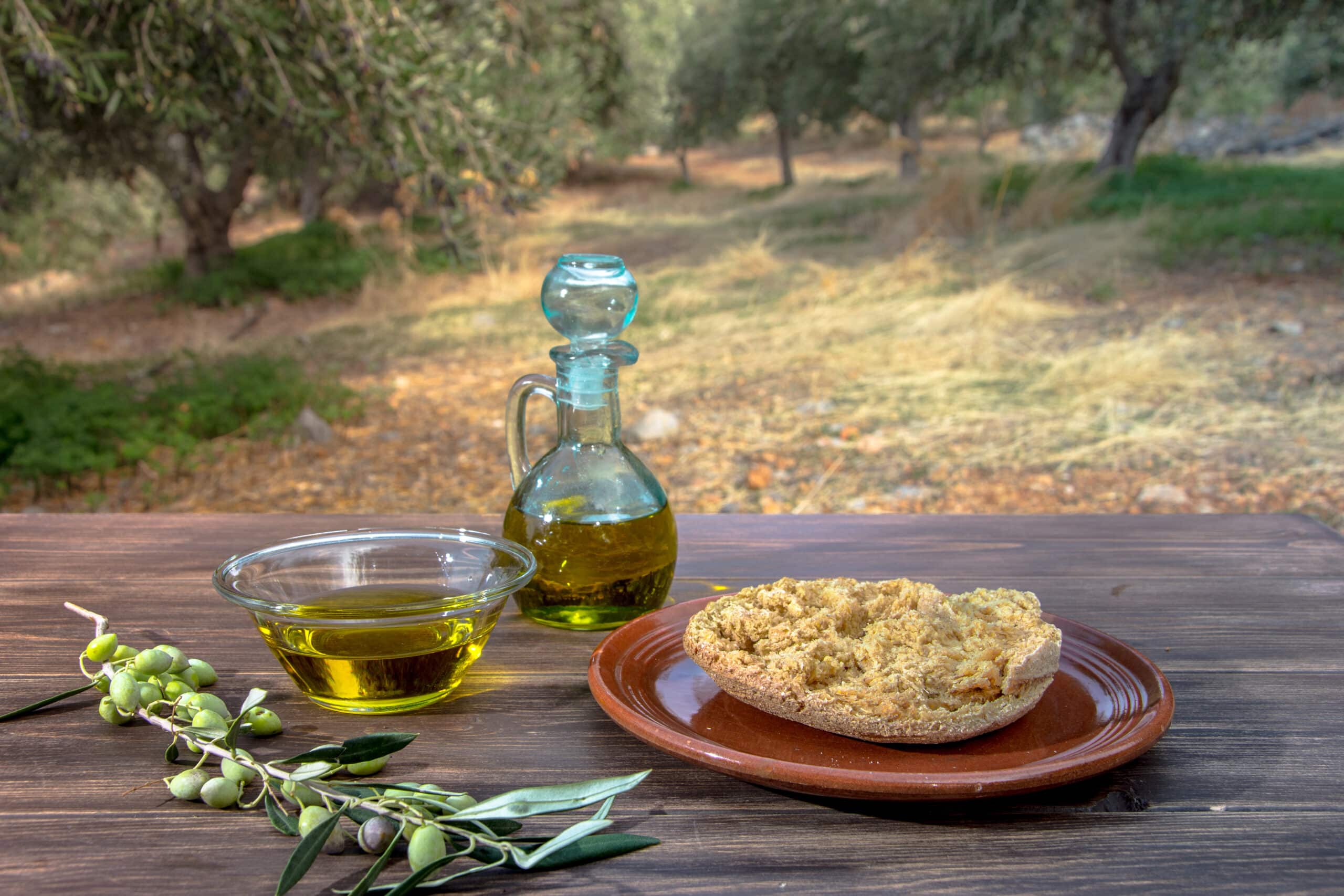 A glass bottle and bowl of olive oil with a piece of bread on a wooden table, set against a blurred background of a Greek olive grove.