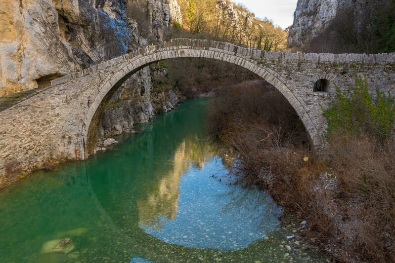 An ancient stone arched bridge spanning a calm, turquoise river in a rocky Greek canyon, perfect for scenic motorcycle touring and adventure travel.